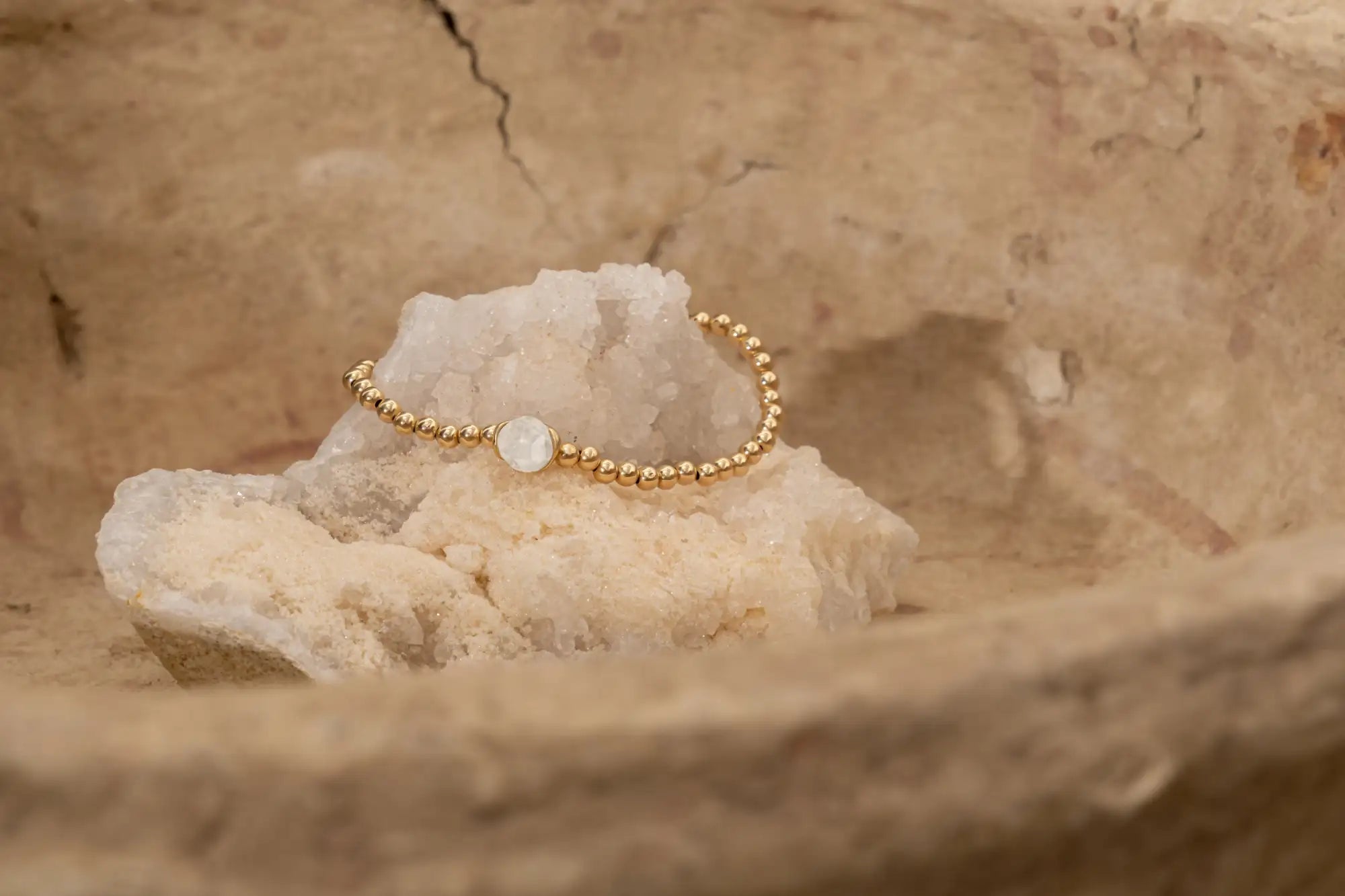 Goudgetinte kralenarmband met een centrale witte maansteenaccent, rustend op kristallijn gesteente.