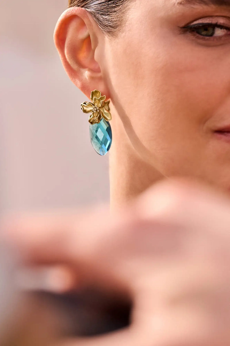 Goudkleurige bloemvormige oorbel met een gefacetteerde blauwe druppelvormige edelsteen die eronder hangt.